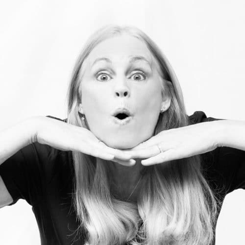 A black and white photo of a blonde woman smiling and framing her face with her hands.