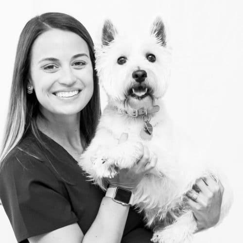 A black and white photo of a woman smiling, wearing scrubs, and holding a cute white dog.