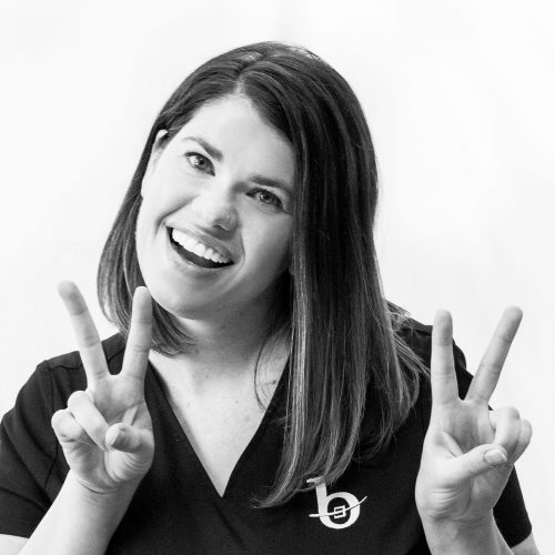 A black and white photo of a woman smiling and making a double peace sign with her hands.