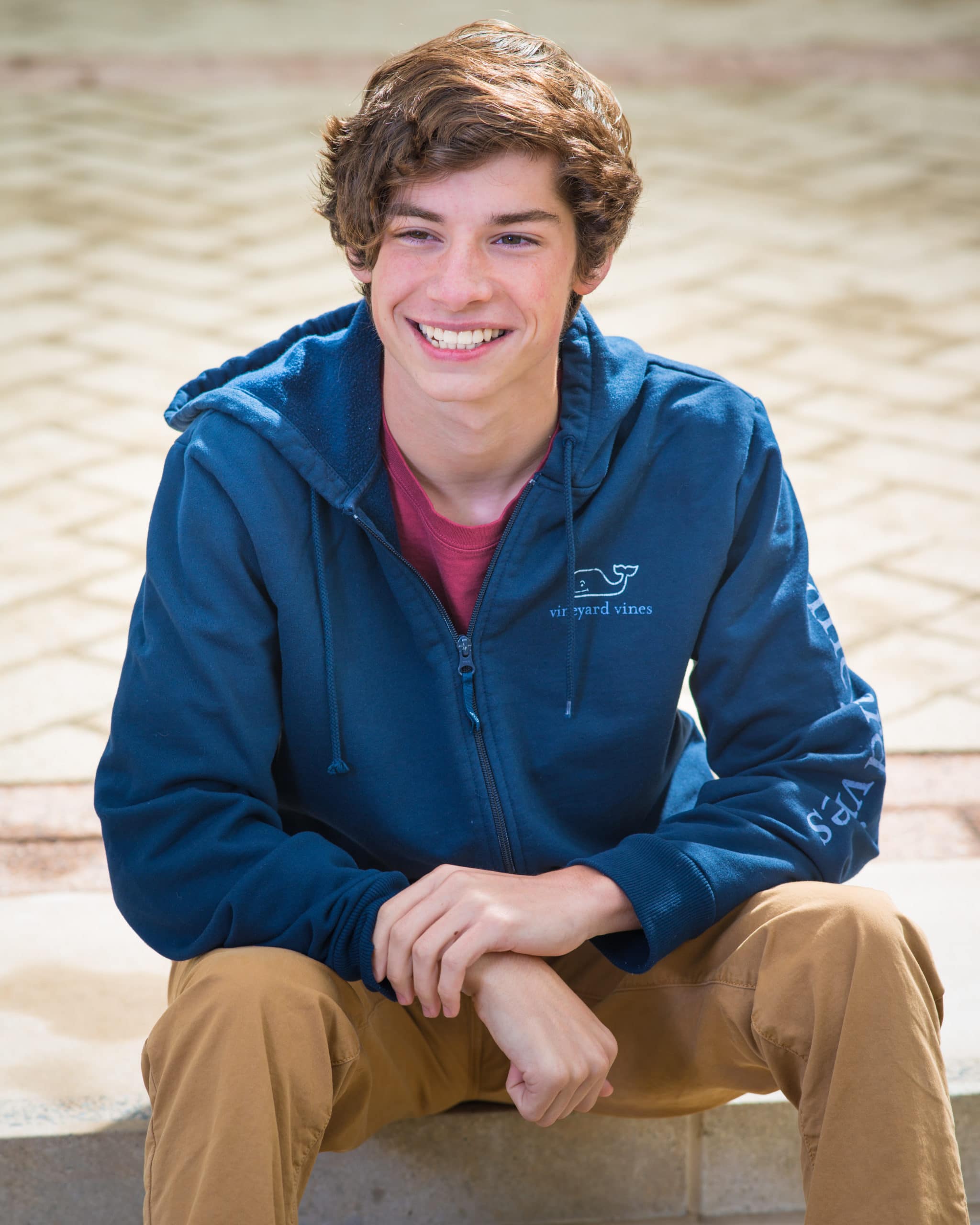A teen boy with brown hair smiling and sitting on stairs. He wears a blue Vineyard Vines sweatshirt.