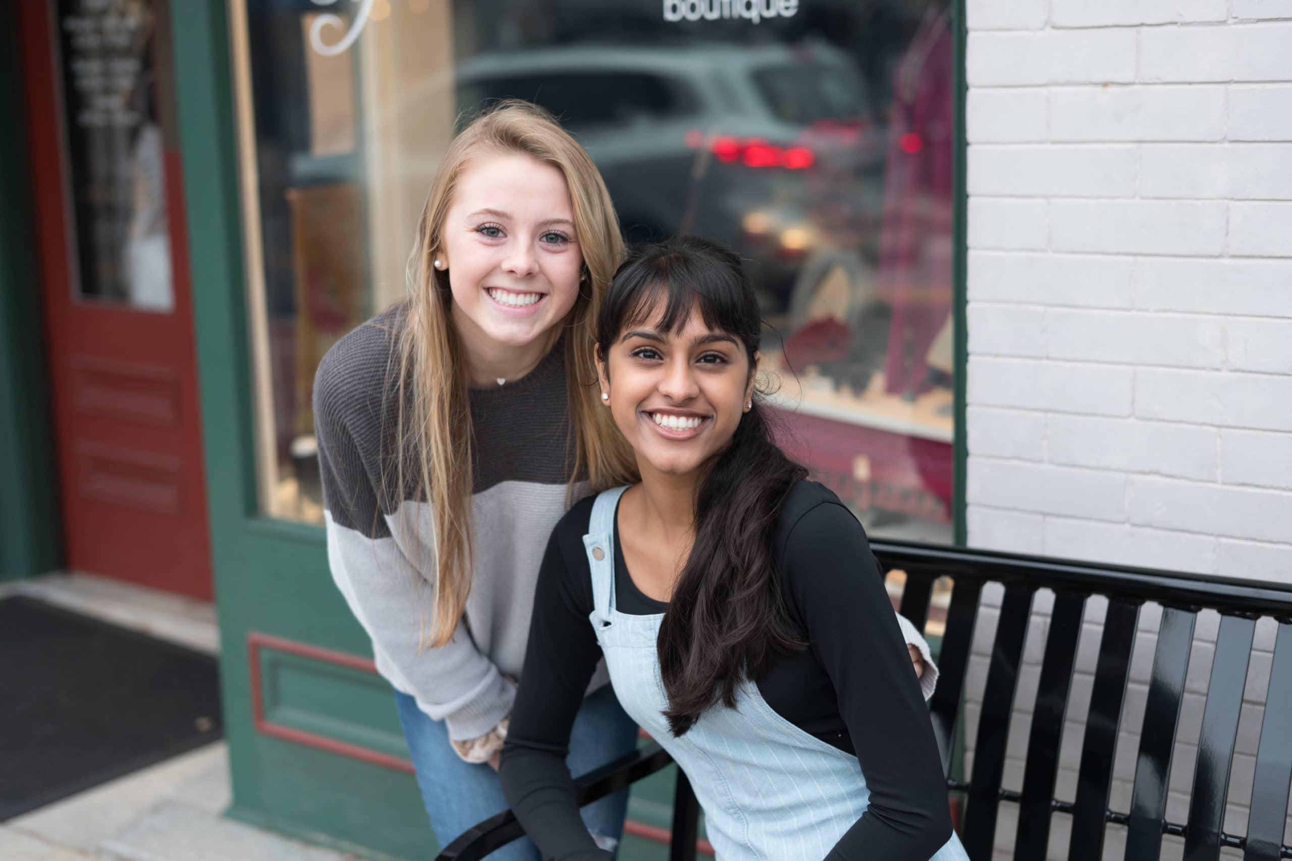 Two teen girls. One is sitting on a bench and one has her arm around the other girl.