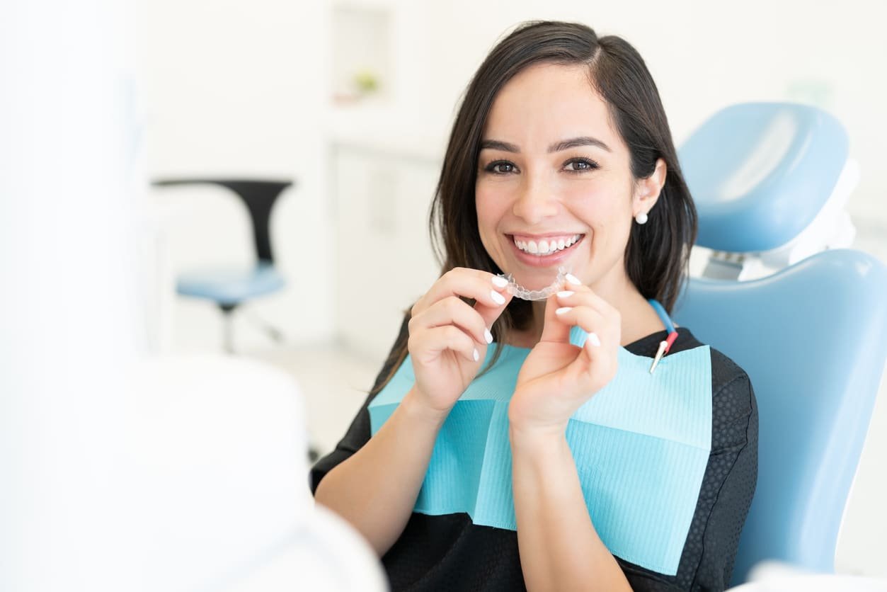 A female patient sitting in a treatment chair holding up a clear aligner tray.