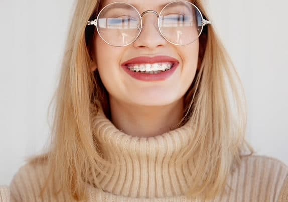 A girl with a turtleneck and glasses smiling with clear braces on.