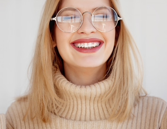 A girl with a turtleneck and glasses smiling with clear braces on.