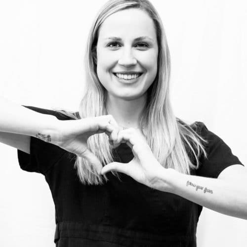 A black and white photo of a blonde woman smiling and making a heart shape with her hands.