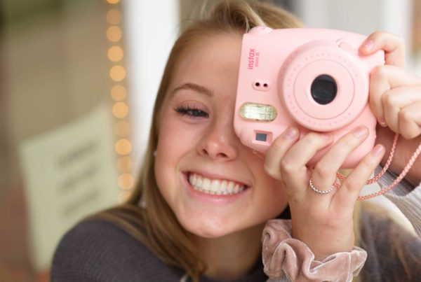 A teen girl with red hair smiling and taking a photo with a Polaroid camera.