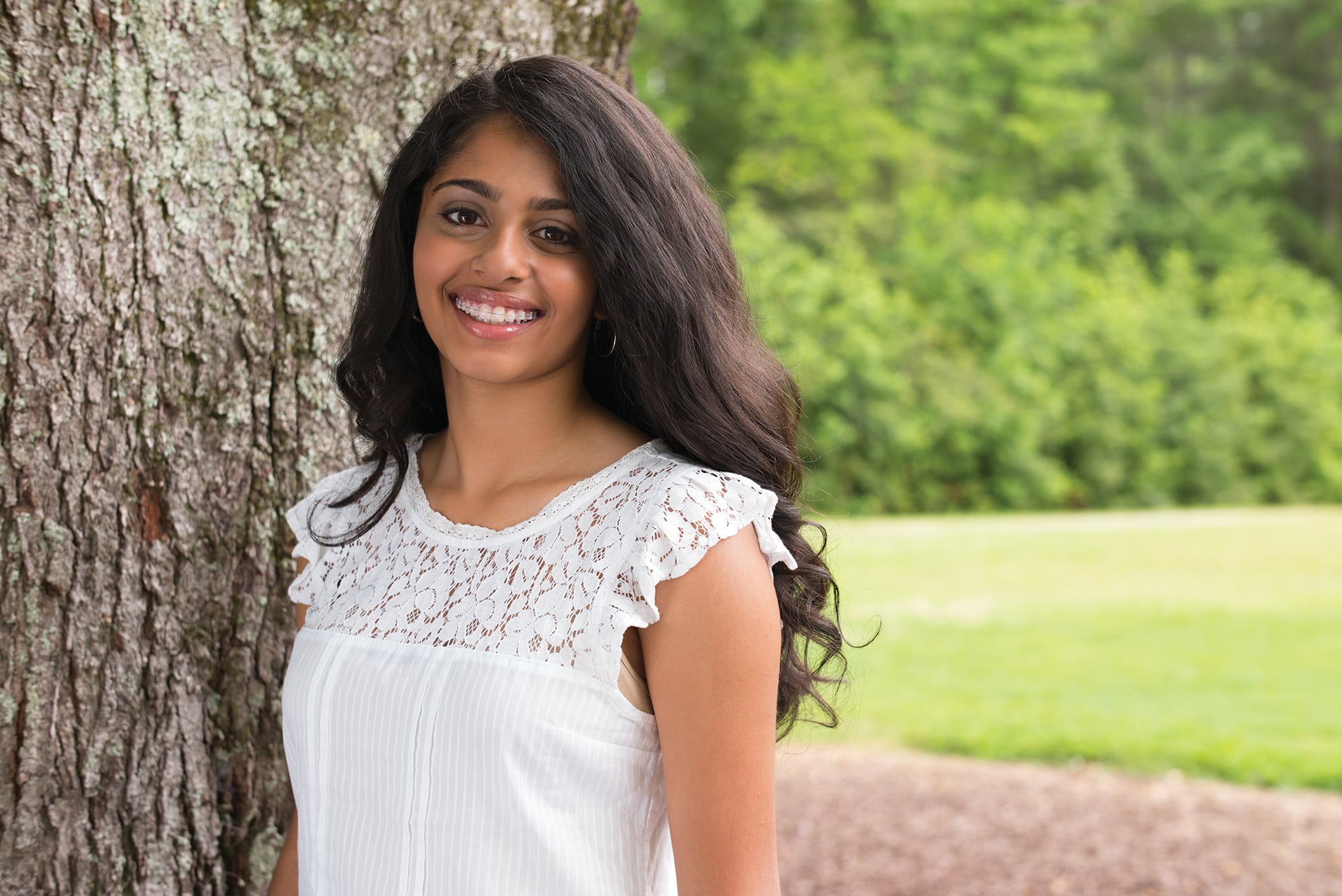 A girl with a white blouse smiling in front of a tree.