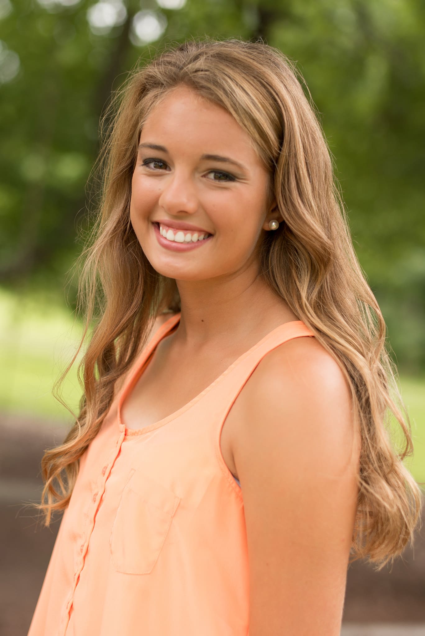 A teen girl with blonde hair and an orange blouse, smiling outdoors.