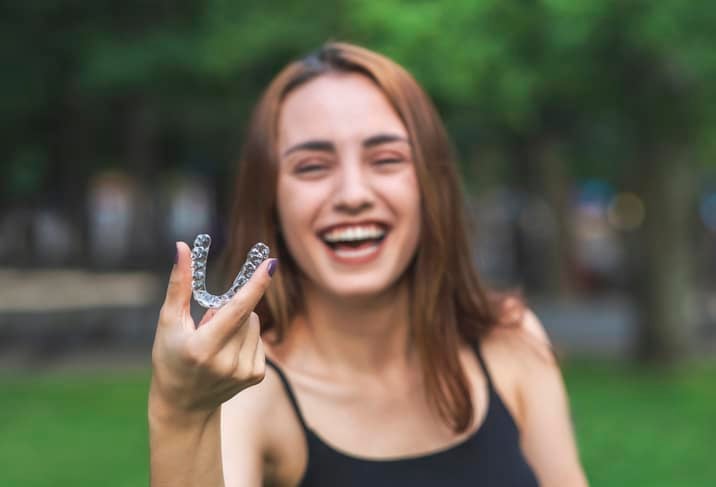 A brown-haired woman holding a clear aligner tray in a park.