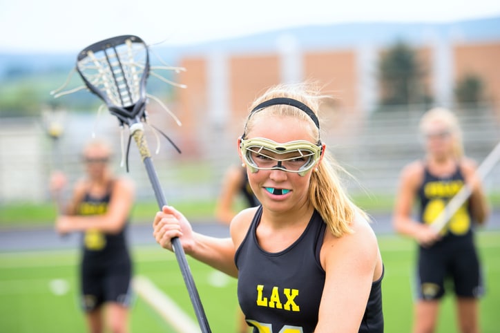 A blonde teen girl wearing a mouthguard while playing lacrosse.