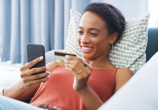 A woman laying on the couch looking at her credit card and phone to make a purchase.