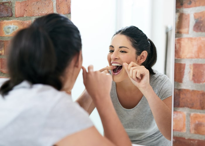 A black-haired woman flossing her teeth while looking in the mirror.