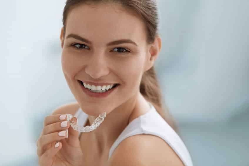 A woman with light brown hair smiling with a clear aligner in her hand.