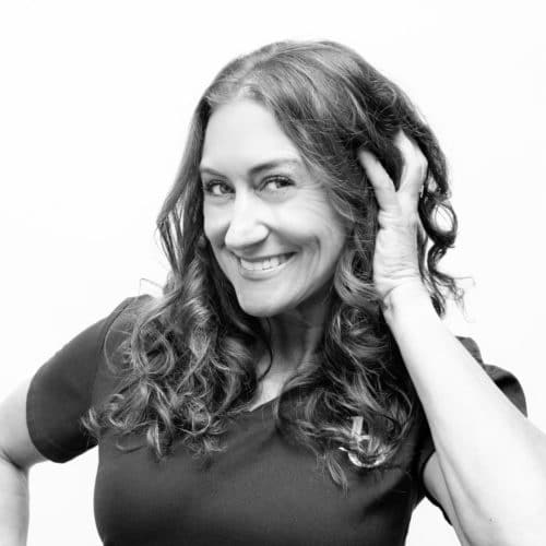 A black and white photo of a curly-haired woman smiling and fluffing her hair with her hand.