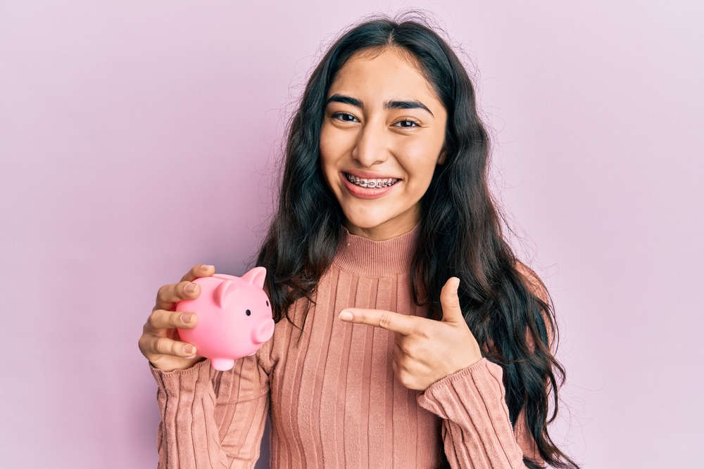 Teen girl with black hair and braces pointing to a piggy bank.