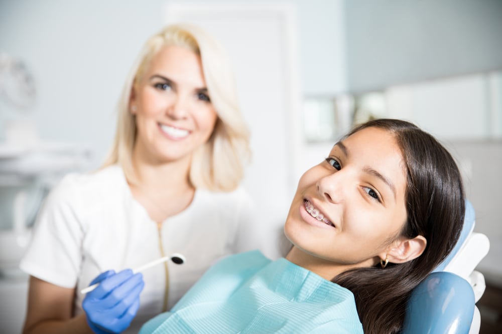 An orthodontist with a dental mirror and a girl with braces in a treatment chair smiling.