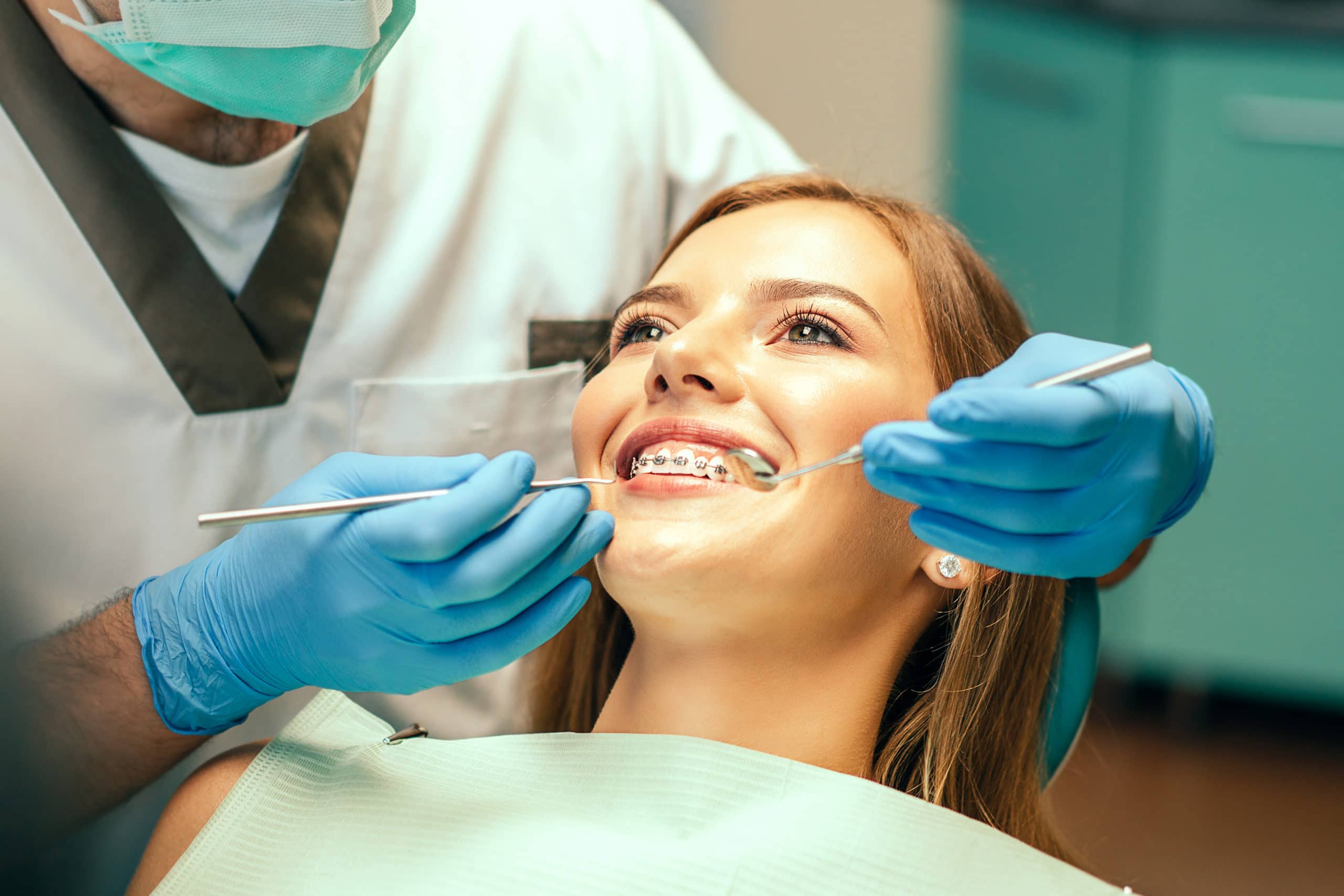 Orthodontist with a mask examining a girl's braces with a dental mirror and a pick.