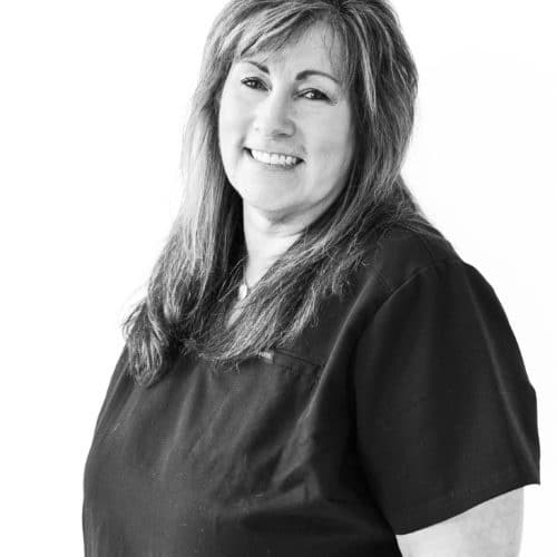 Black and white portrait of a woman wearing black dental scrubs while smiling at the camera.